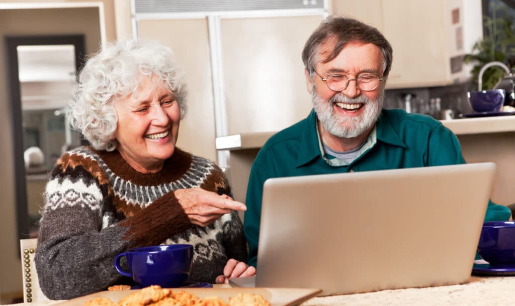 Senior couple smiling and laughing, sitting in their kitchen and looking at a laptop.