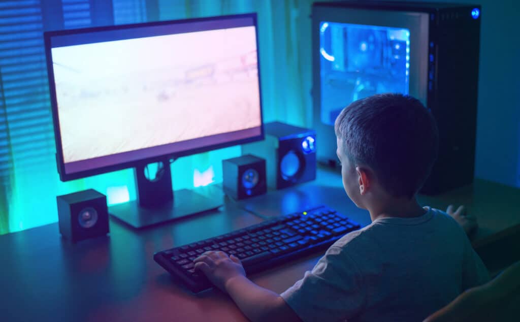 A young boy playing video games on a desktop computer in a dark room lit with neon lights