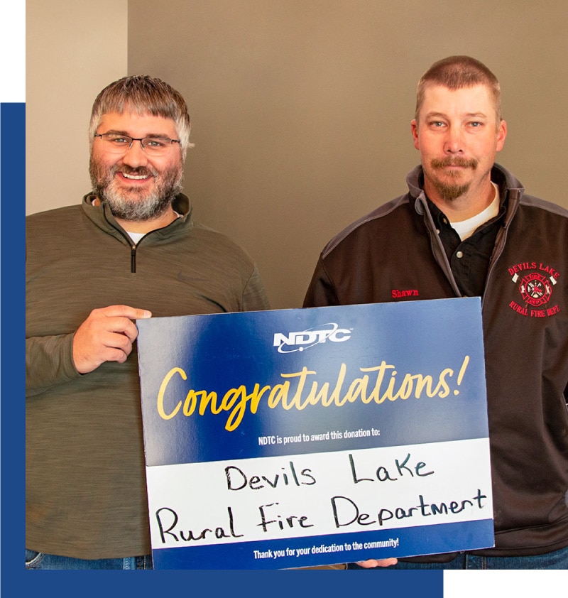 A photo of a male NDTC employee awarding Dollars in Motion Funds to a male representative of the Devils Lake Fire Department